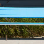 A modern blue 970 bench with metal legs stands on a concrete surface in front of tall grass and a dark building wall.