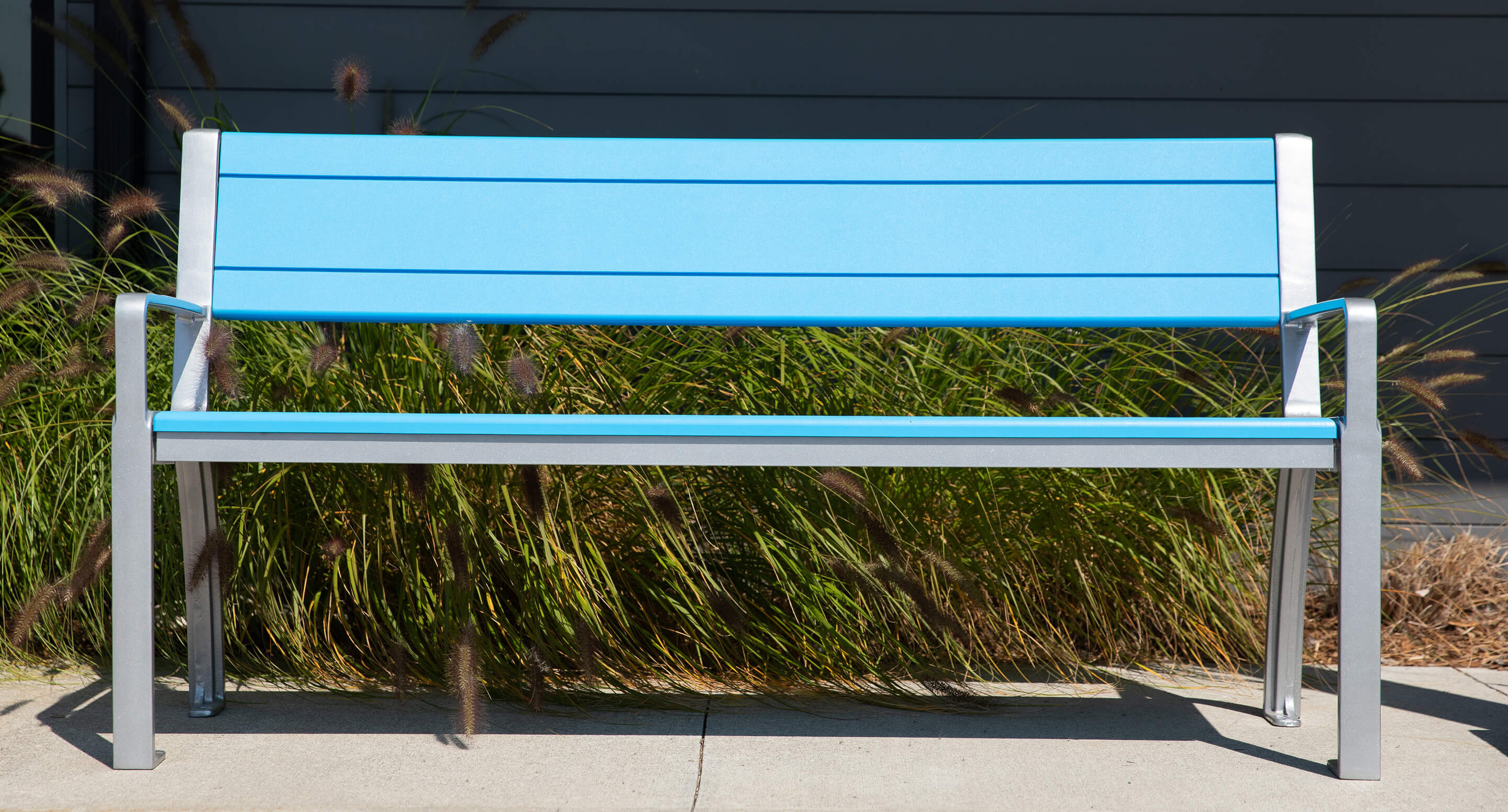 A modern blue 970 bench with metal legs stands on a concrete surface in front of tall grass and a dark building wall.