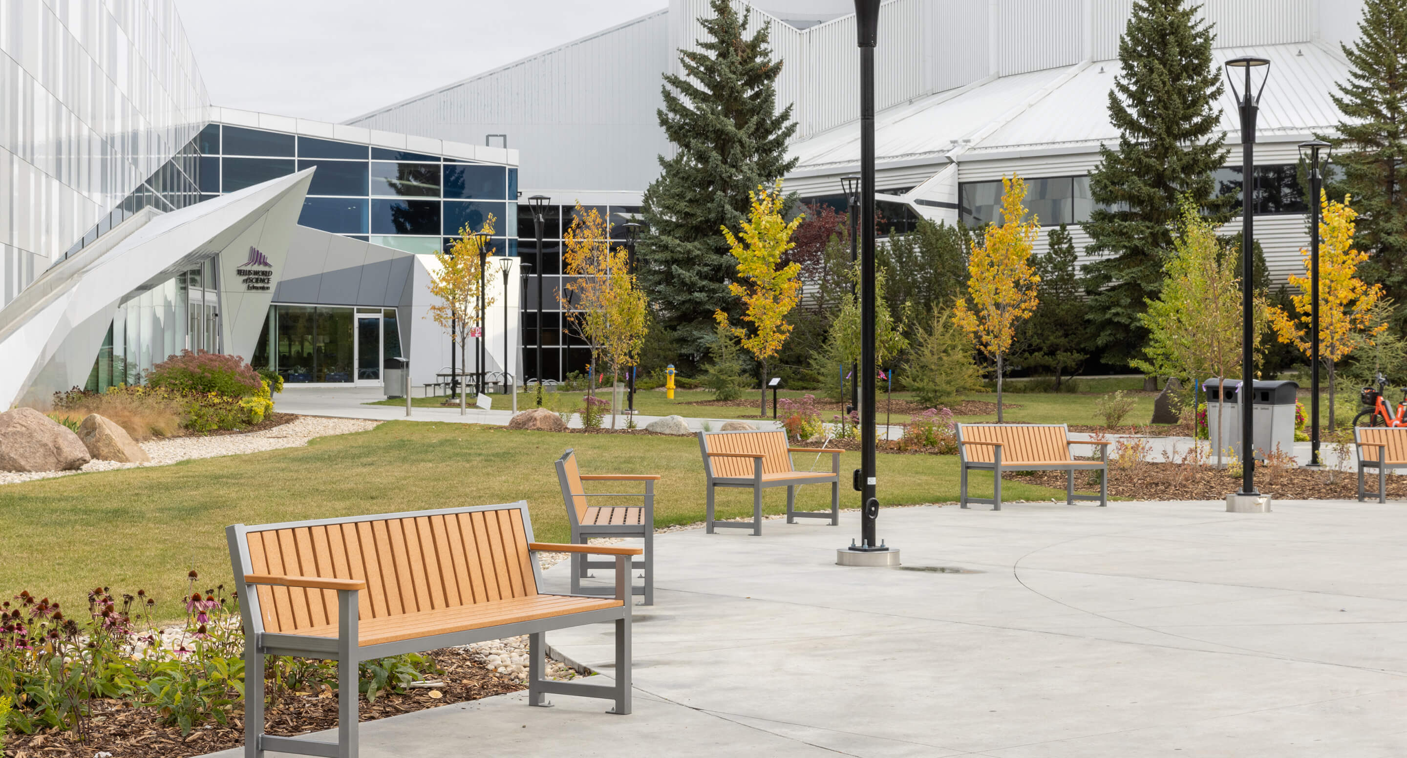 maglin-benches-400-telus-world Modern outdoor courtyard with several wooden benches, trees with autumn foliage, lamp posts, and a glass building entrance in the background.