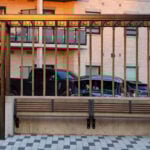 Wooden bench with a slatted pergola above, located in a paved urban courtyard with flowers, parked cars, and apartments in the background.