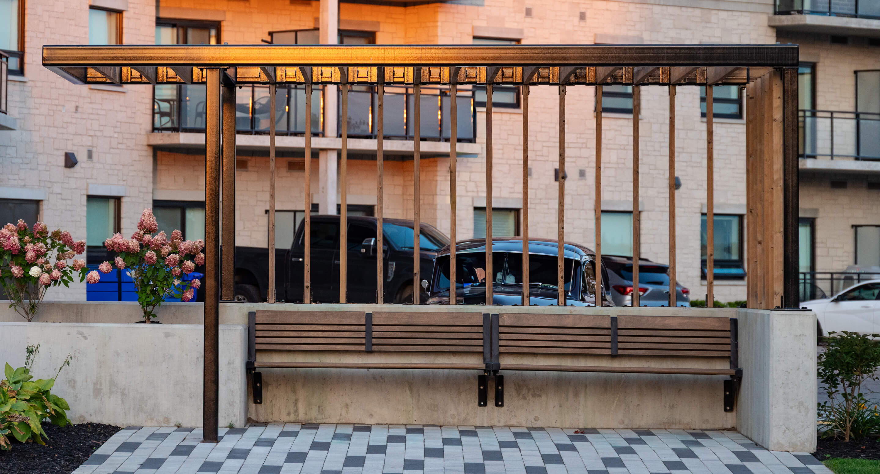 Wooden bench with a slatted pergola above, located in a paved urban courtyard with flowers, parked cars, and apartments in the background.