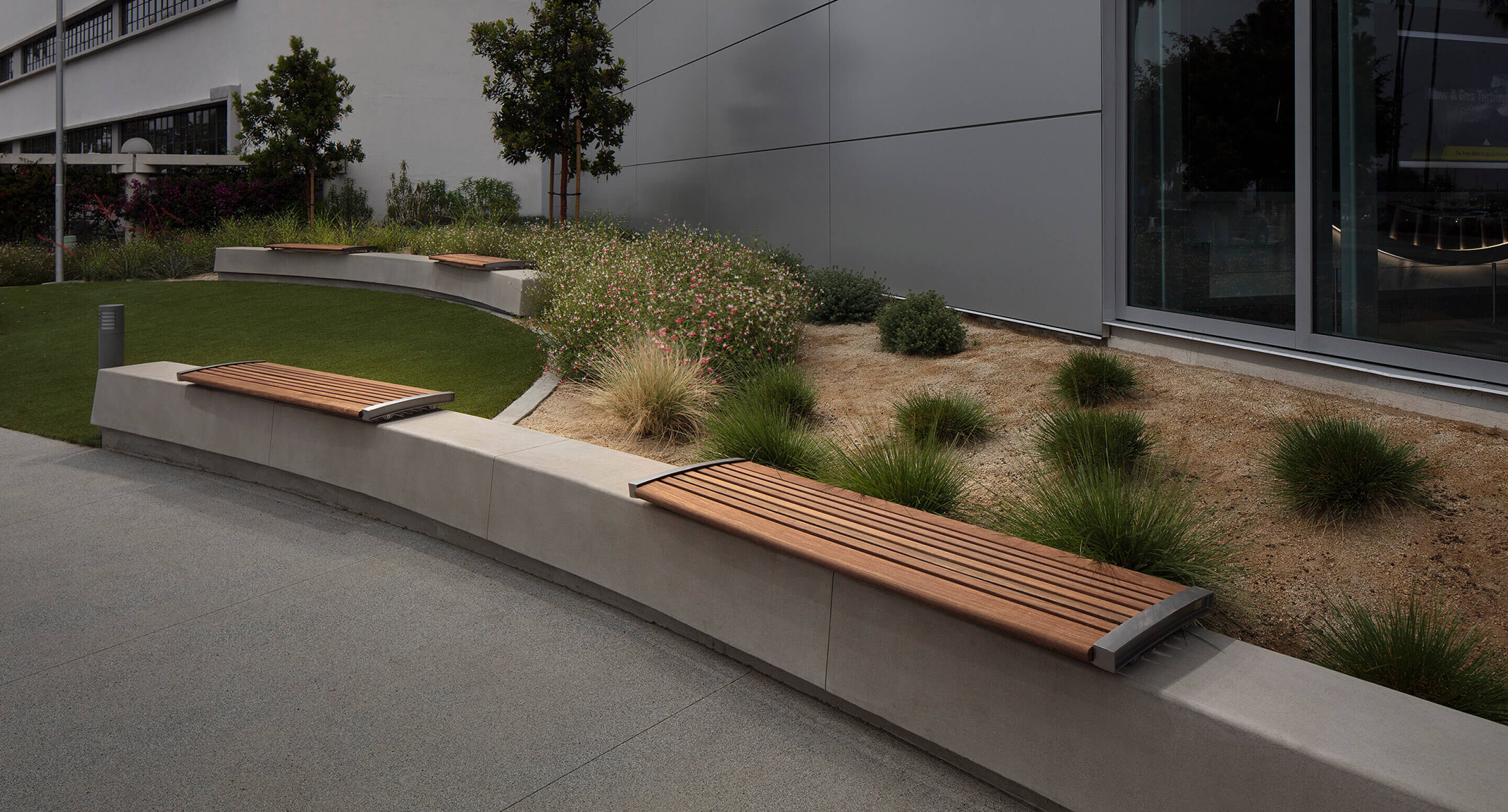 Curved concrete with backless 720 wall-mount benches in a landscaped outdoor area next to a modern building, surrounded by grass, shrubs, and small trees.
