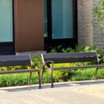 Two empty metal 870 benches are placed side by side on a sidewalk in front of a modern building with large windows and landscaping.