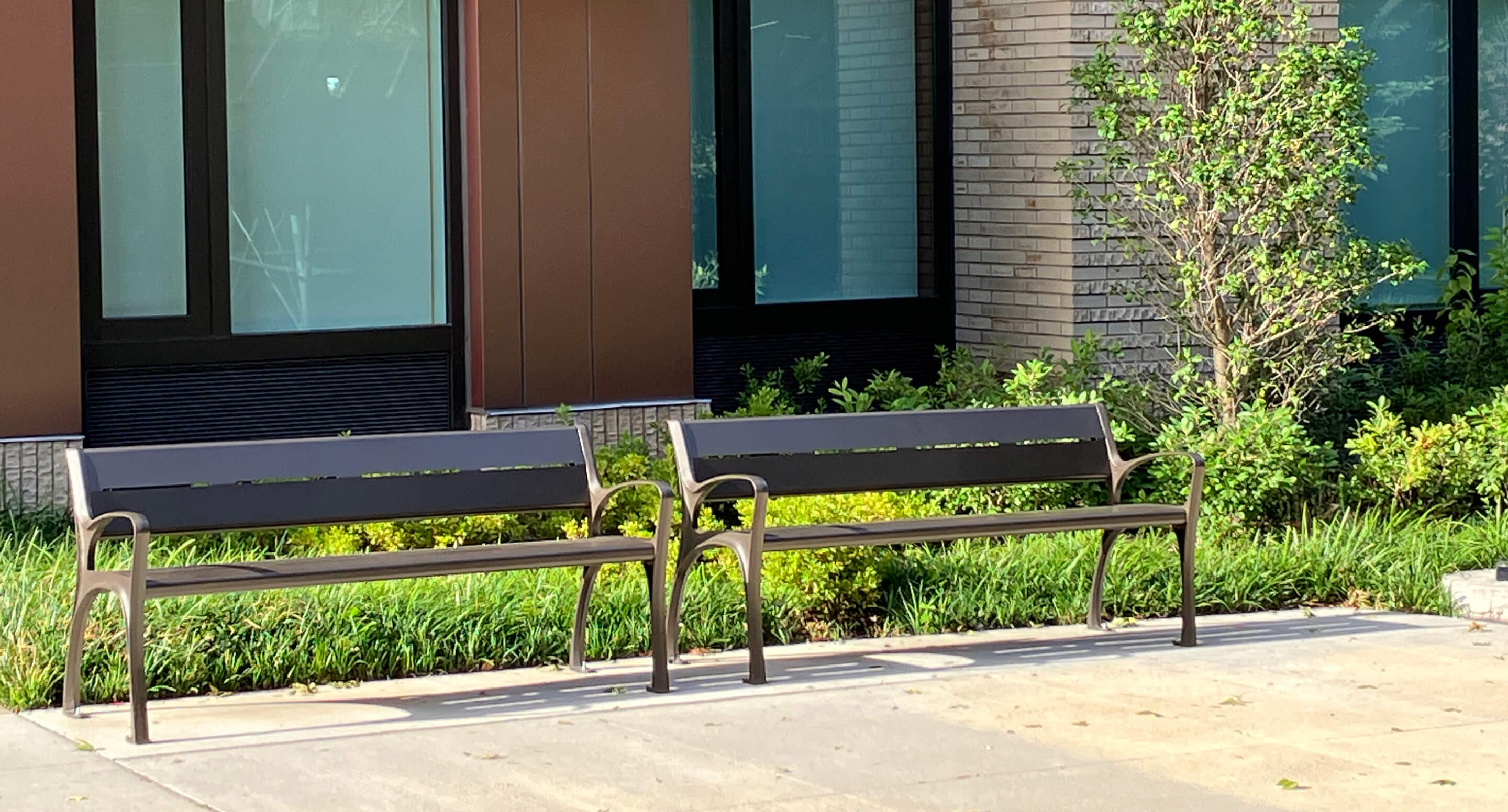 maglin-benches-870-metal Two empty metal 870 benches are placed side by side on a sidewalk in front of a modern building with large windows and landscaping.
