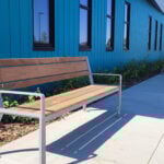 A wooden bench with metal arms is placed on a sidewalk next to a blue building with several windows and landscaping.