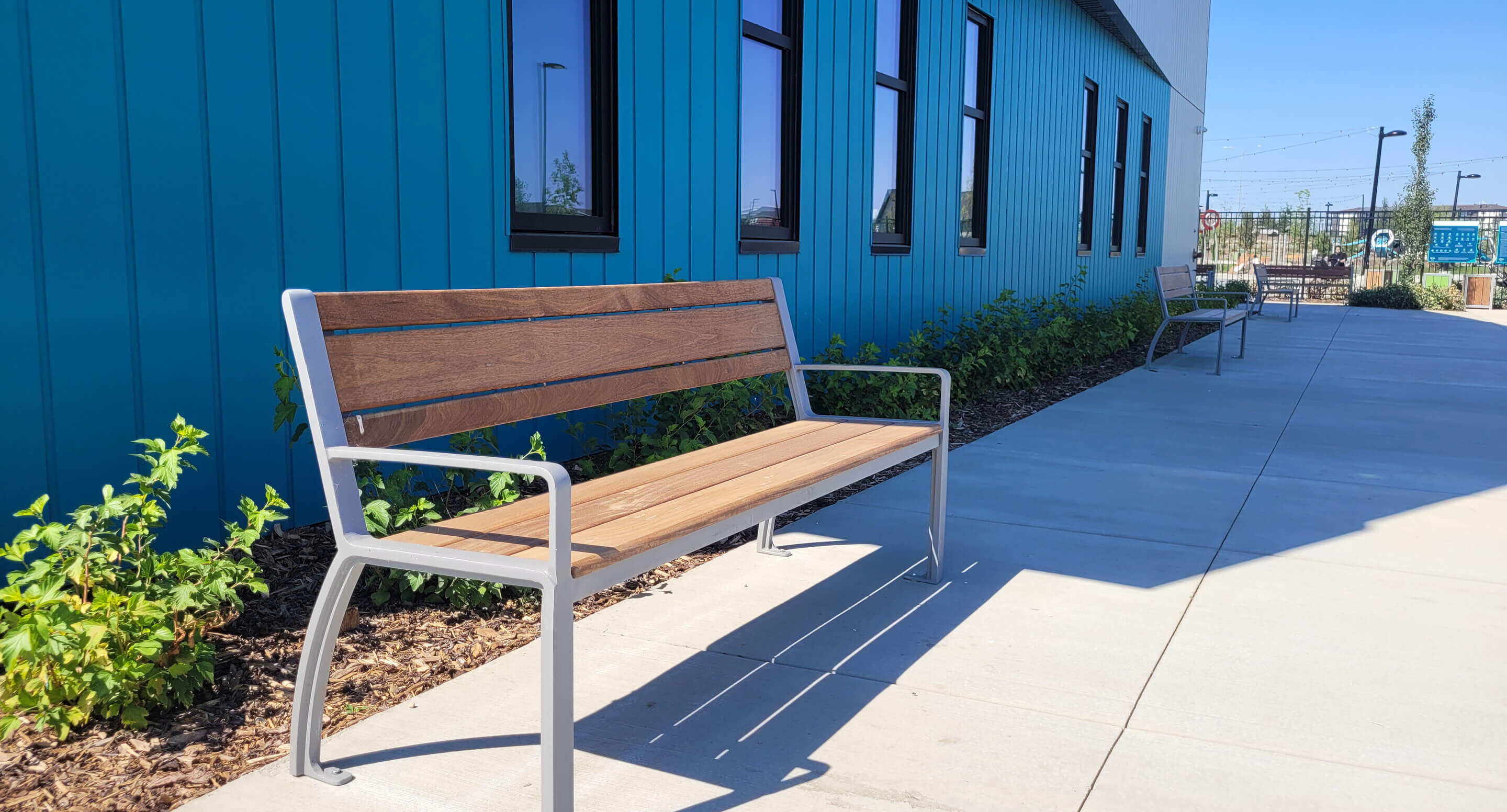 A wooden bench with metal arms is placed on a sidewalk next to a blue building with several windows and landscaping.