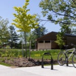 A bicycle is parked at a bike rack on a sunny day, with a wooden building and green trees in the background.