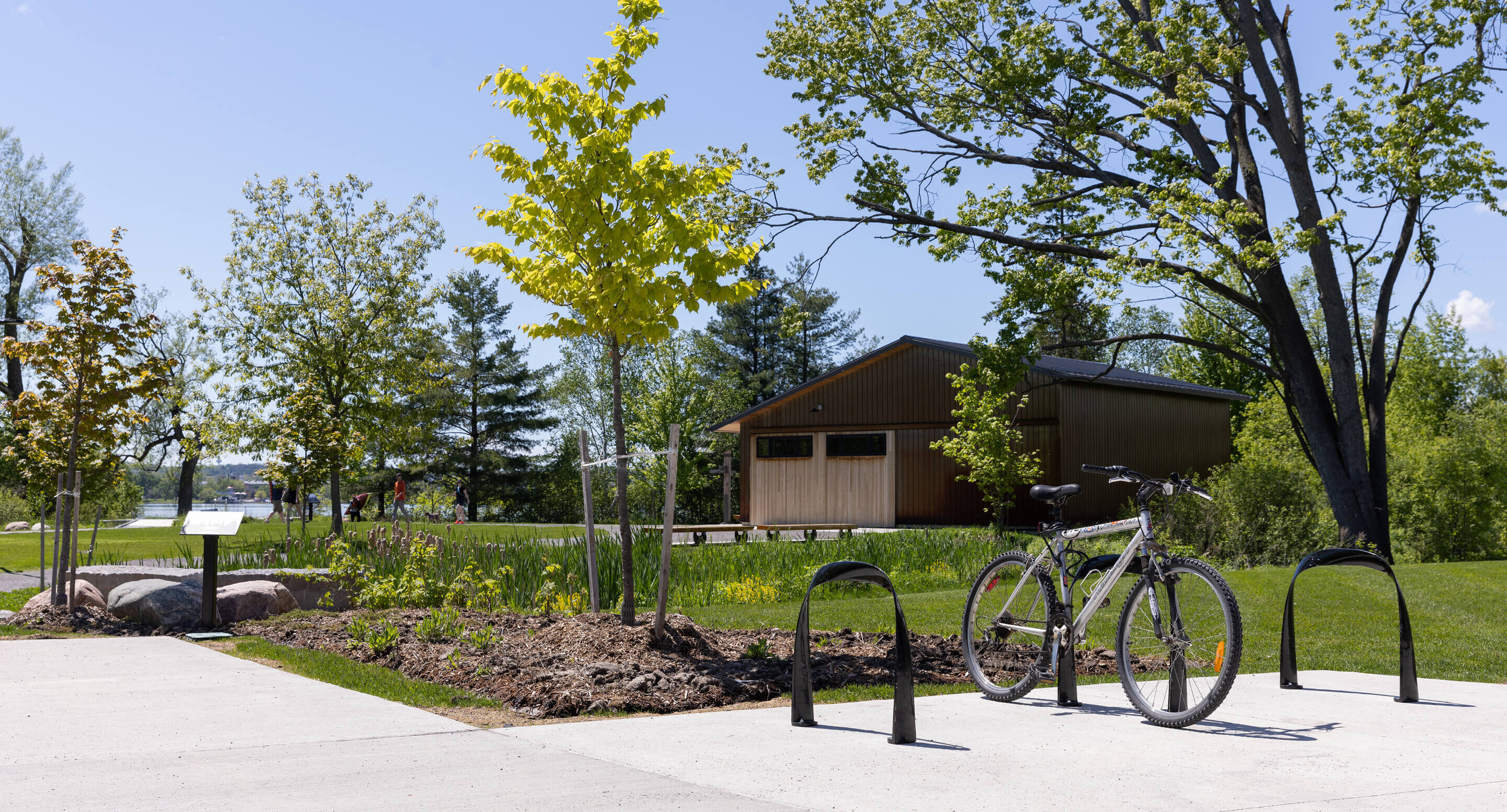 A bicycle is parked at a bike rack on a sunny day, with a wooden building and green trees in the background.