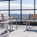 Rooftop patio with two wooden benches and a chair beside a decorative fire pit filled with white stones, overlooking a cityscape through glass panels.