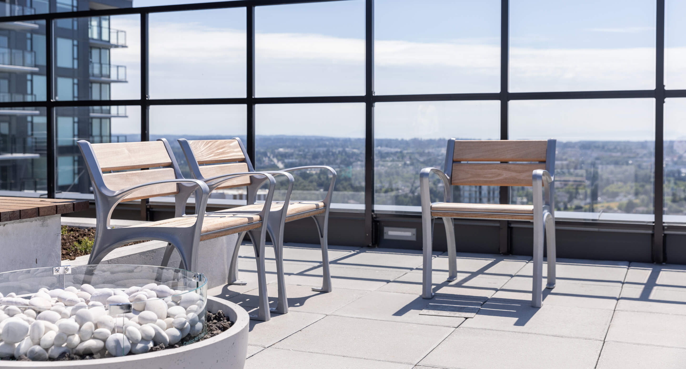 maglin-chairs-870-king-george Rooftop patio with two wooden benches and a chair beside a decorative fire pit filled with white stones, overlooking a cityscape through glass panels.