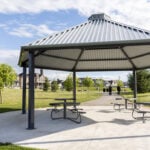 A metal gazebo with picnic tables sits on a concrete pad in a grassy park, with houses and a few trees visible in the background under a partly cloudy sky.
