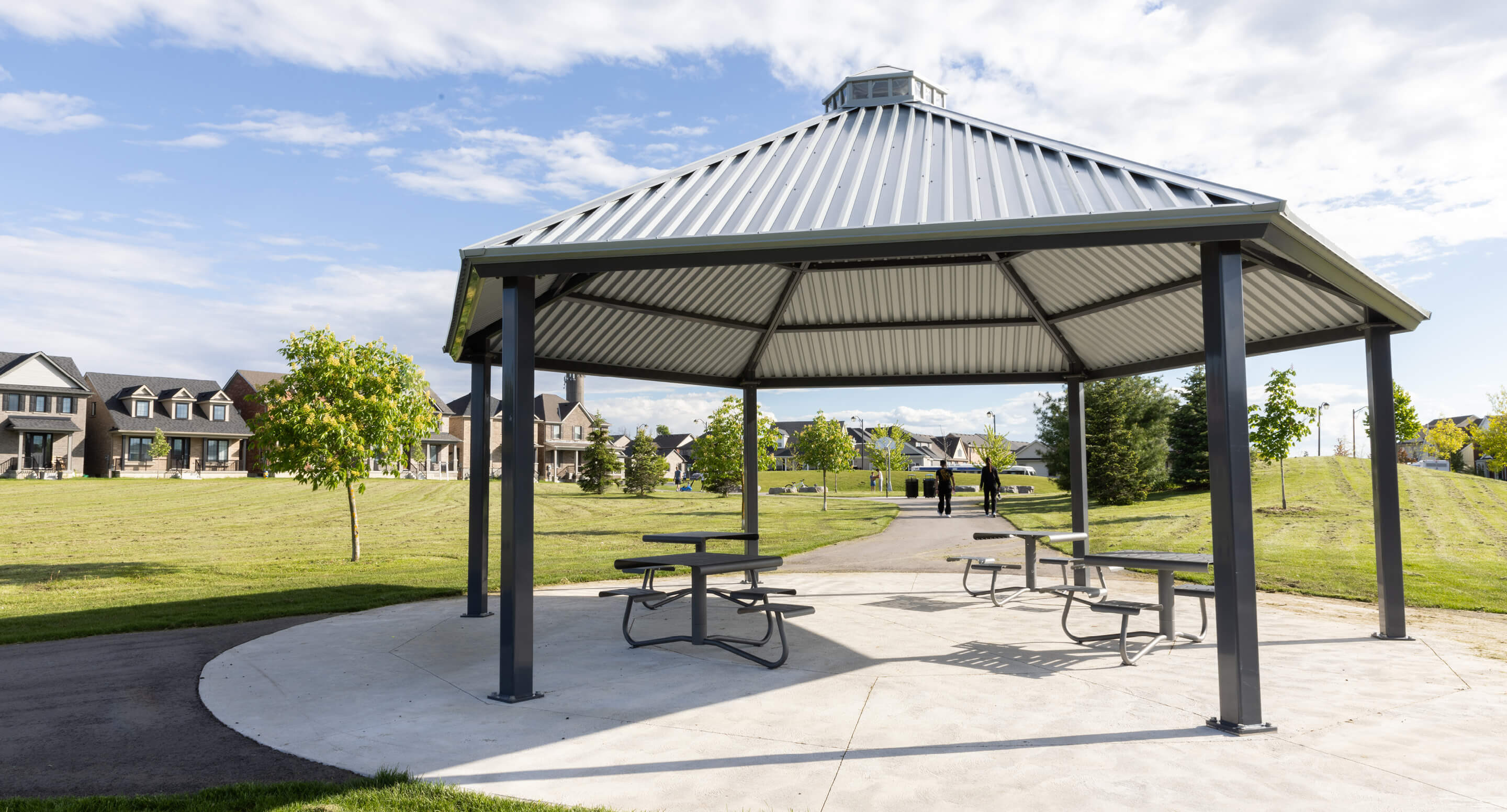 A metal gazebo with picnic tables sits on a concrete pad in a grassy park, with houses and a few trees visible in the background under a partly cloudy sky.
