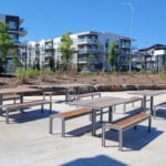 Several picnic tables and benches are arranged on a concrete area near apartment buildings, with landscaping and trees in the background under a clear blue sky.