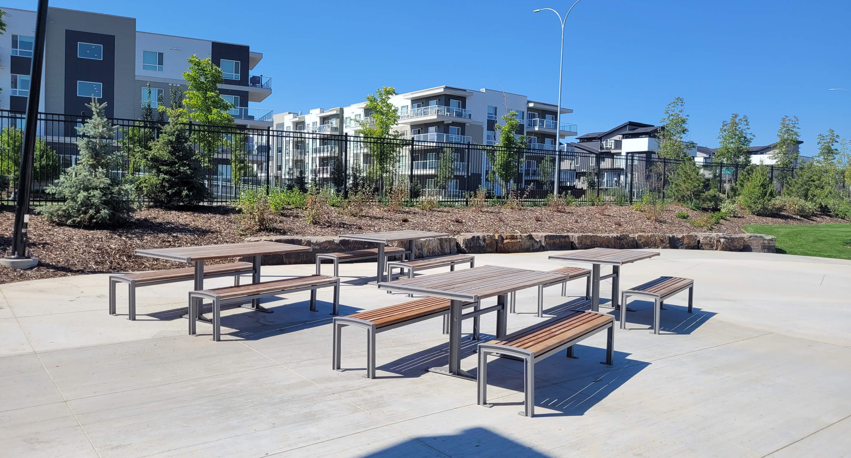Several picnic tables and benches are arranged on a concrete area near apartment buildings, with landscaping and trees in the background under a clear blue sky.