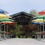 Several picnic tables with colorful, flower-shaped umbrellas are arranged under a pavilion in an outdoor park setting with trees in the background.