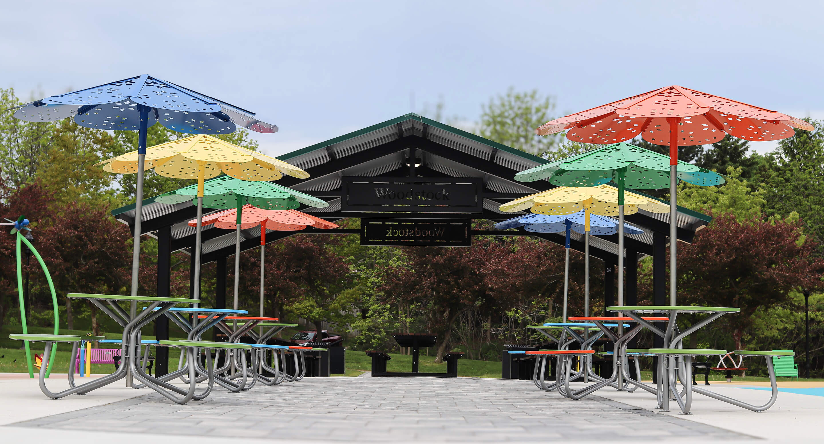 Several picnic tables with colorful, flower-shaped umbrellas are arranged under a pavilion in an outdoor park setting with trees in the background.