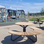 Circular wooden picnic tables are placed on a concrete area near a playground with climbing structures, slides, and nearby houses under a blue sky.