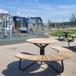 Circular wooden picnic tables are placed on a concrete area near a playground with climbing structures, slides, and nearby houses under a blue sky.