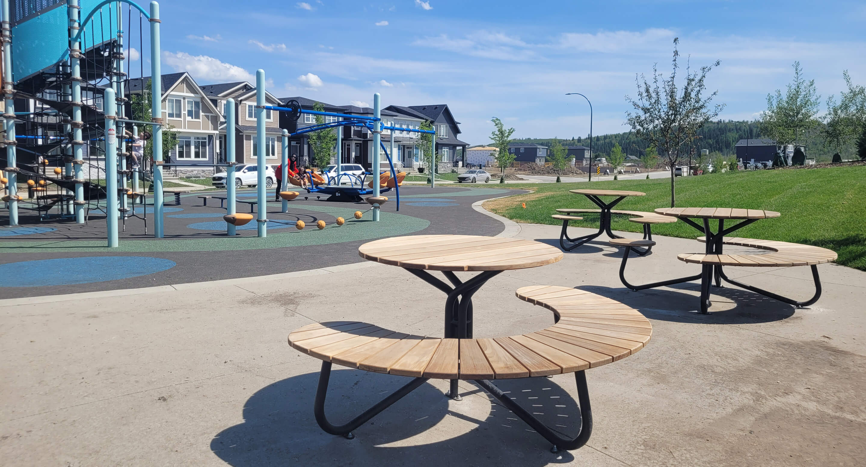 Circular wooden picnic tables are placed on a concrete area near a playground with climbing structures, slides, and nearby houses under a blue sky.