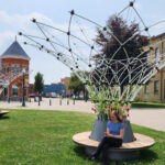 A person sits on a circular wooden bench around a modern metal and flower structure in a grassy area with historic brick buildings in the background.