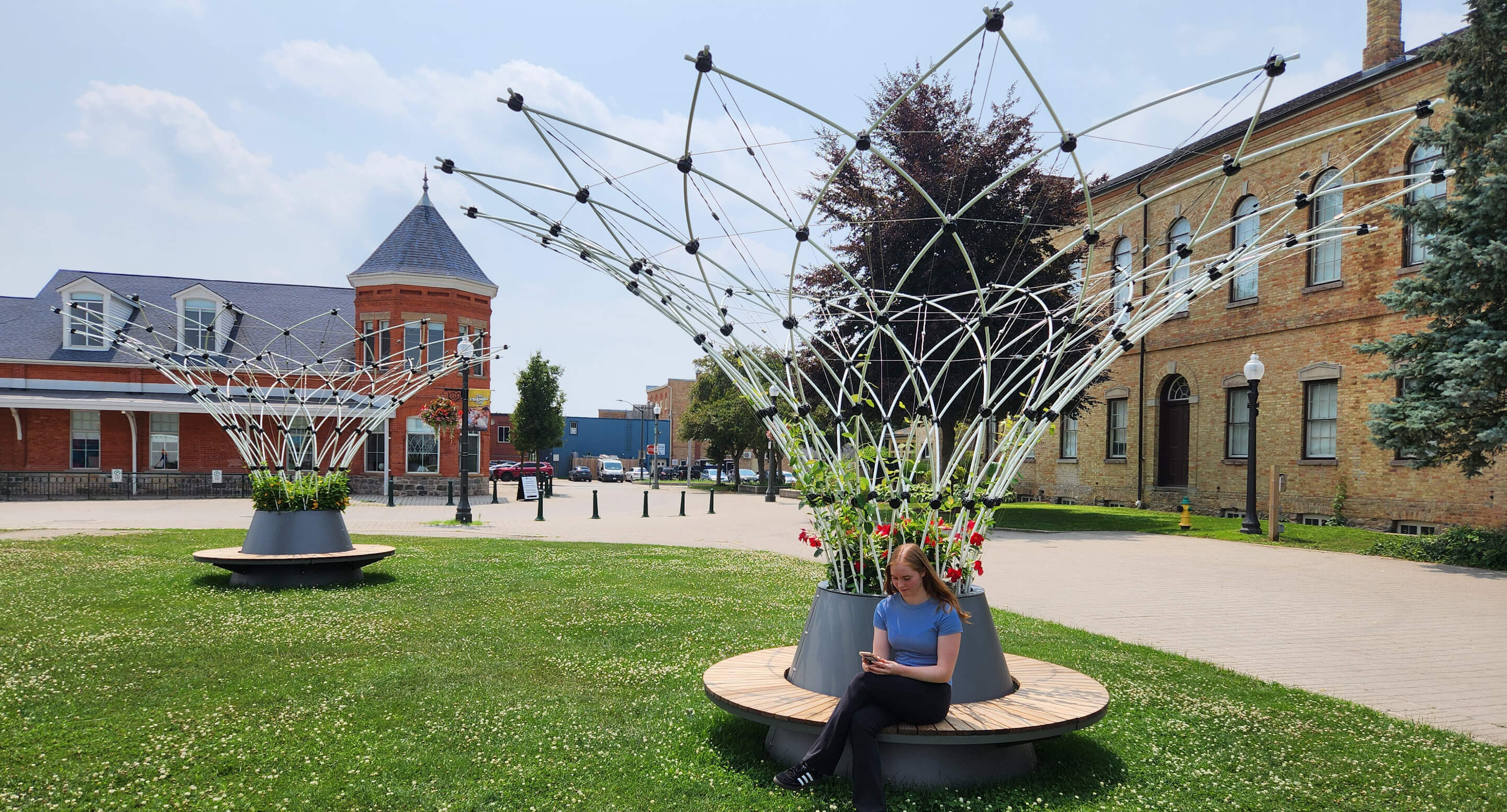 A person sits on a circular wooden bench around a modern metal and flower structure in a grassy area with historic brick buildings in the background.