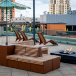 Outdoor patio with wooden benches, sun loungers, a fire pit, and an umbrella, set on a tiled terrace with city buildings in the background.