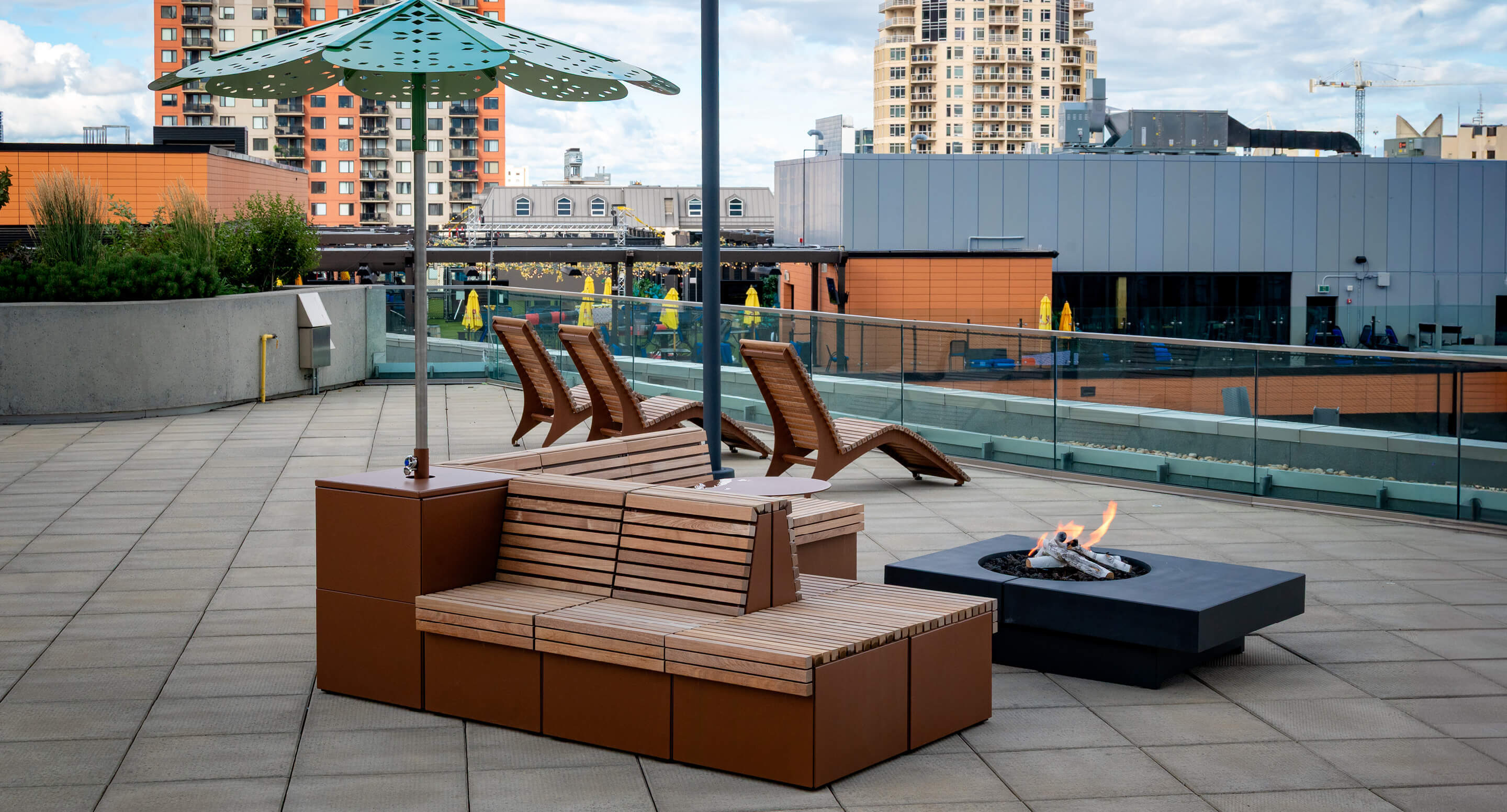 maglin-pixel-hero-img-1 Outdoor patio with wooden benches, sun loungers, a fire pit, and an umbrella, set on a tiled terrace with city buildings in the background.