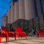 Several bright red chairs are arranged on a paved plaza near large silos and a stone wall, with people nearby and city buildings visible in the background at dusk.