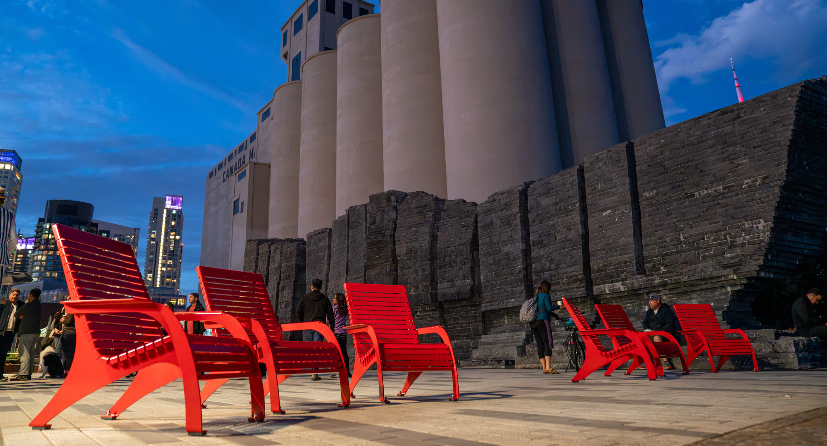 Several bright red chairs are arranged on a paved plaza near large silos and a stone wall, with people nearby and city buildings visible in the background at dusk.