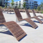 Four modern wooden lounge chairs are arranged in a row on a concrete patio, with apartment buildings and greenery visible in the background.
