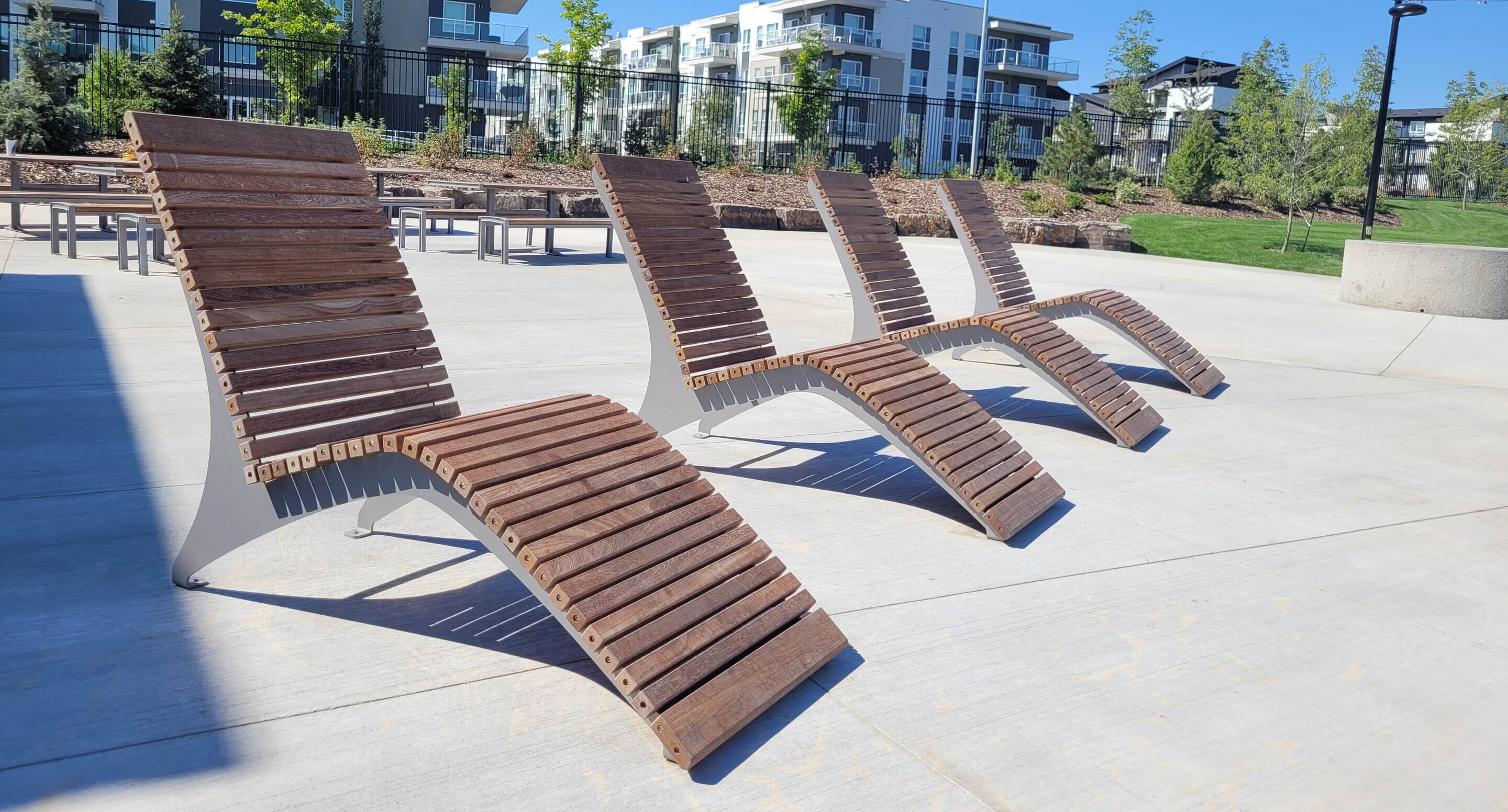 Four modern wooden lounge chairs are arranged in a row on a concrete patio, with apartment buildings and greenery visible in the background.