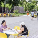 People sit at yellow tables and chairs in an outdoor plaza, talking, eating, and walking, surrounded by trees and greenery on a sunny day.