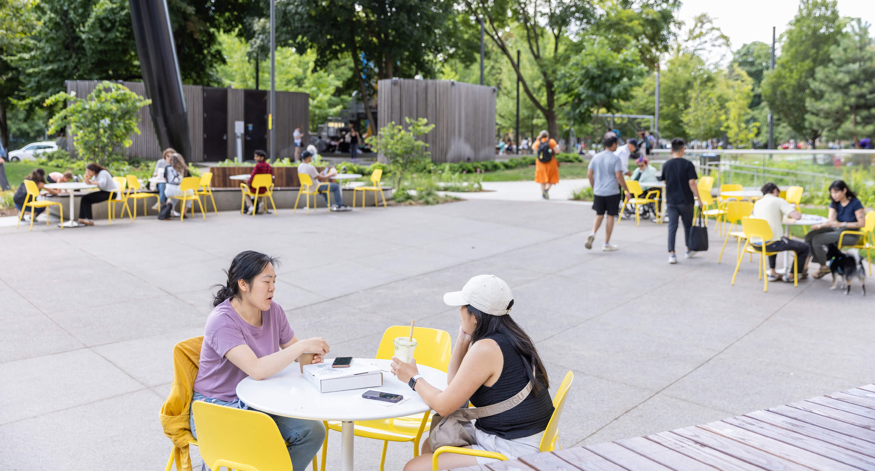 maglin-tables-chairs-aries-battery-ocad-1 People sit at yellow tables and chairs in an outdoor plaza, talking, eating, and walking, surrounded by trees and greenery on a sunny day.