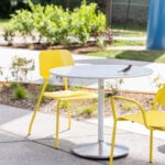 A small bird is perched on a round white table surrounded by three yellow chairs in an outdoor seating area.