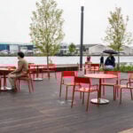 People sit at red tables and chairs on an outdoor wooden deck near a waterfront, with a few trees and industrial buildings in the background.