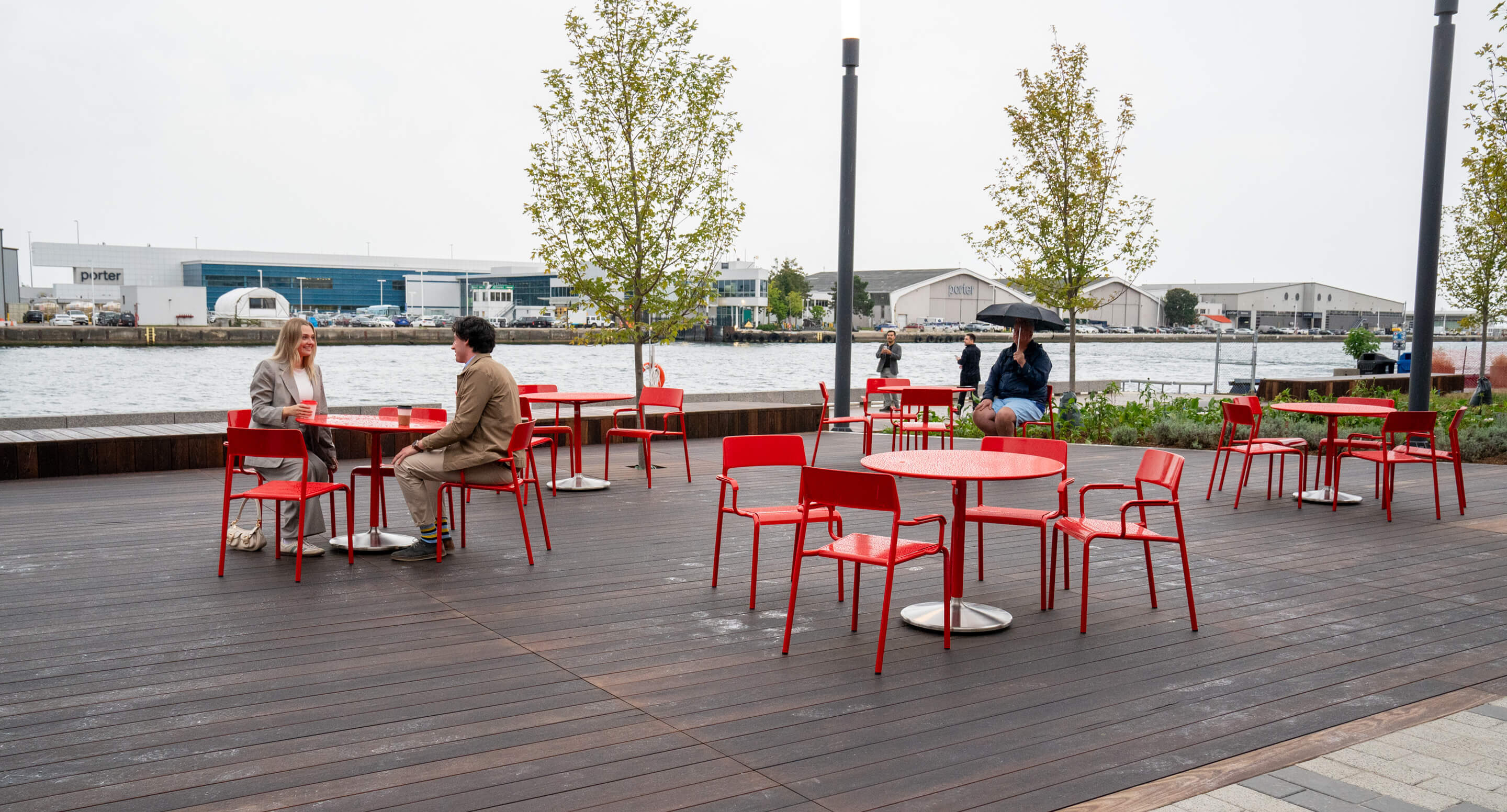 People sit at red tables and chairs on an outdoor wooden deck near a waterfront, with a few trees and industrial buildings in the background.