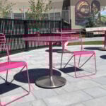 Three pink metal tables with matching chairs are arranged on a paved outdoor patio, with a mural depicting faces on the wall in the background.