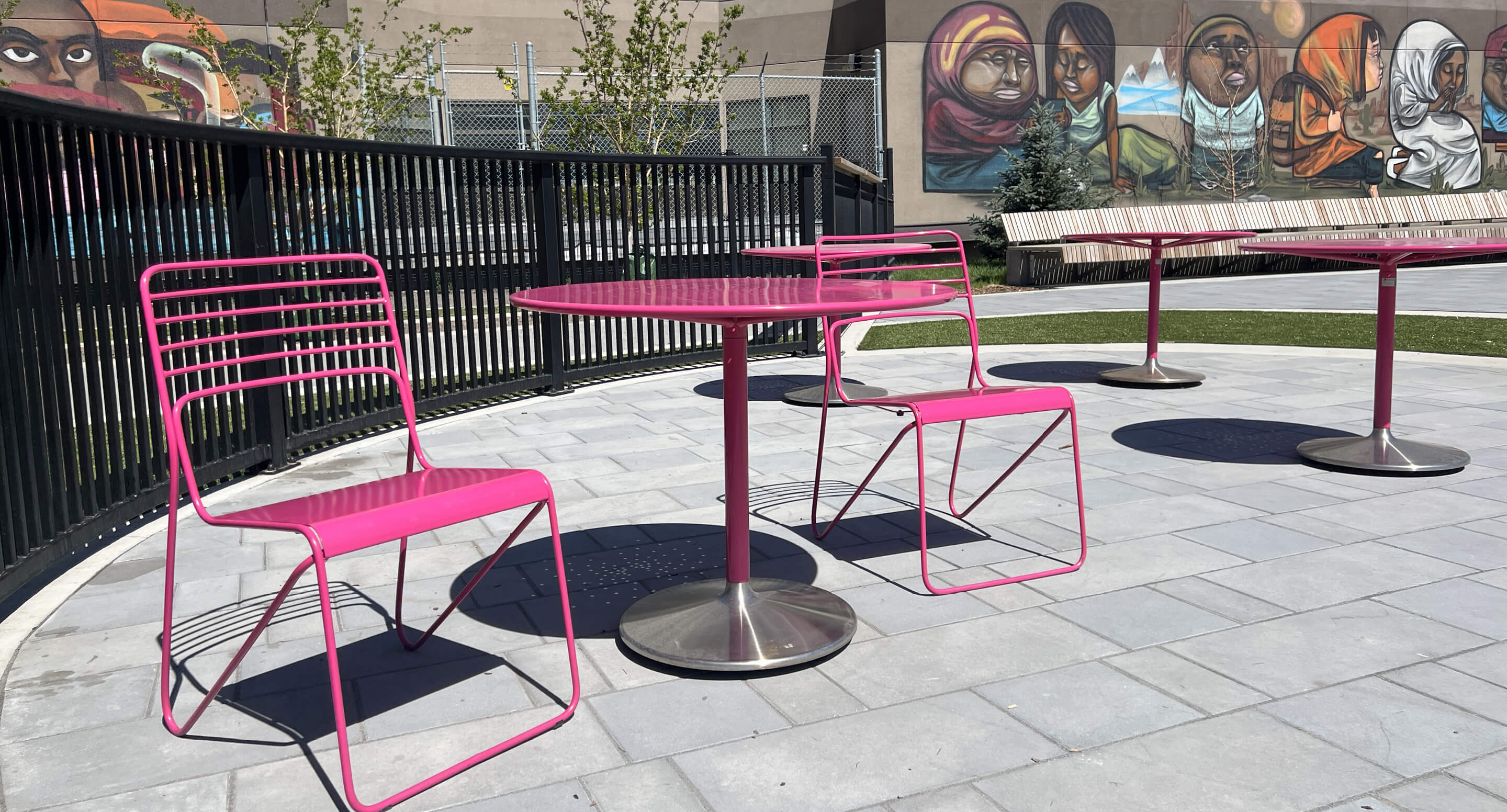 Three pink metal tables with matching chairs are arranged on a paved outdoor patio, with a mural depicting faces on the wall in the background.