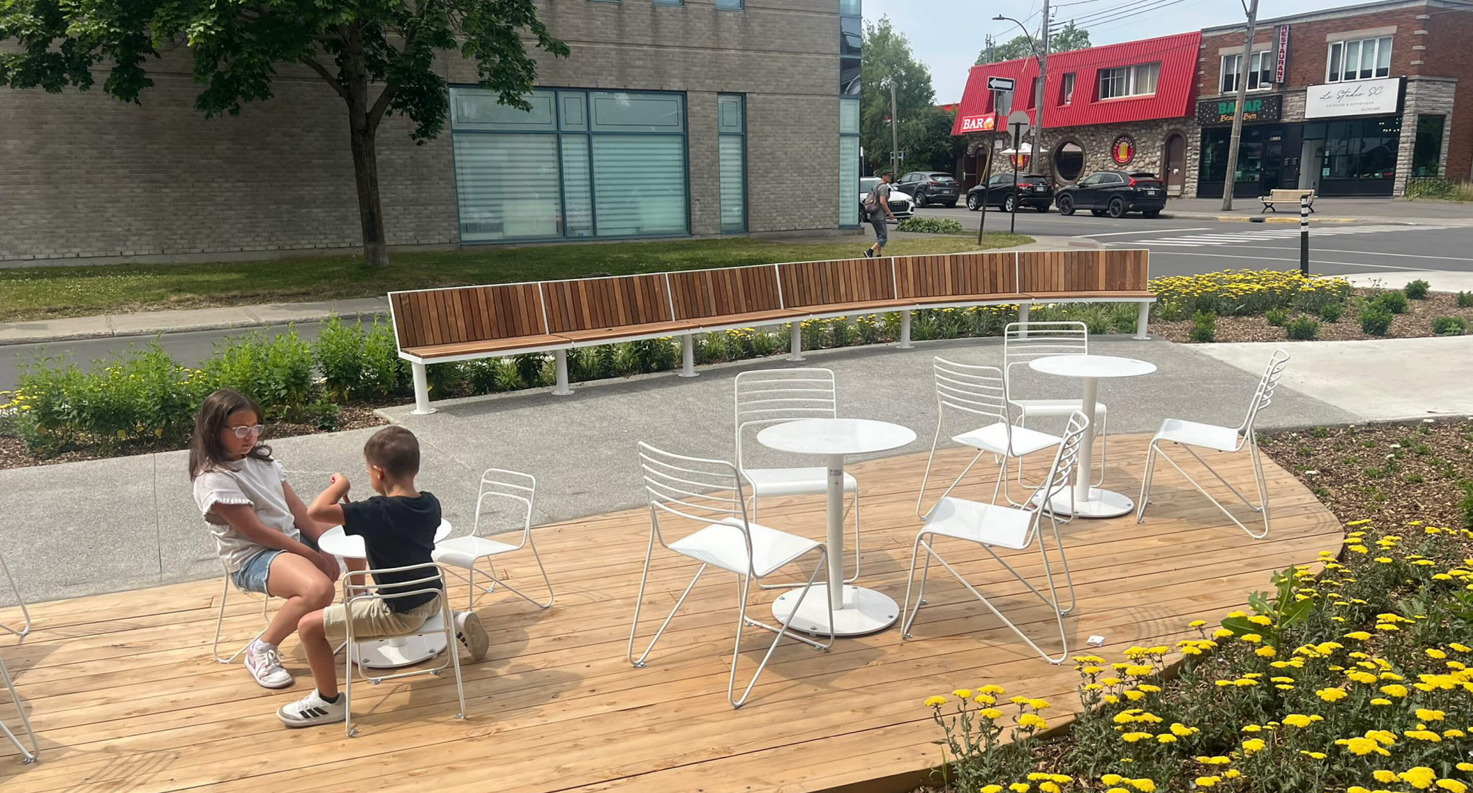 Two children sit at a white table on a wooden deck with empty chairs around them, near a street lined with buildings and yellow flowers in the foreground.