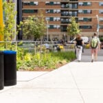 Two people walk along a sidewalk in an urban park area with trash and recycling bins, greenery, and apartment buildings in the background.