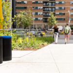 Two people walk along a sidewalk in an urban park area with trash and recycling bins, greenery, and apartment buildings in the background.