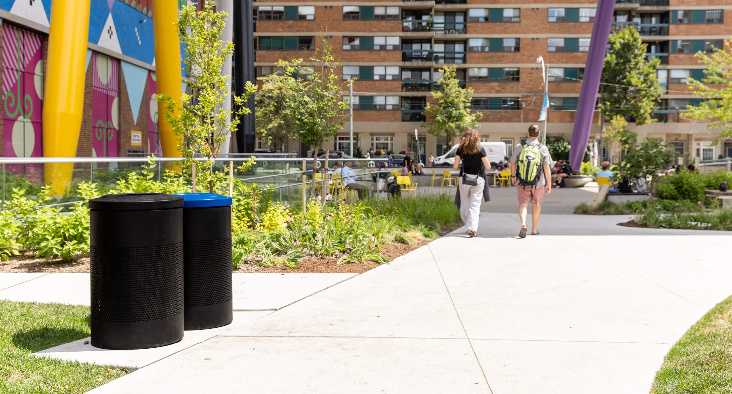 Two people walk along a sidewalk in an urban park area with trash and recycling bins, greenery, and apartment buildings in the background.