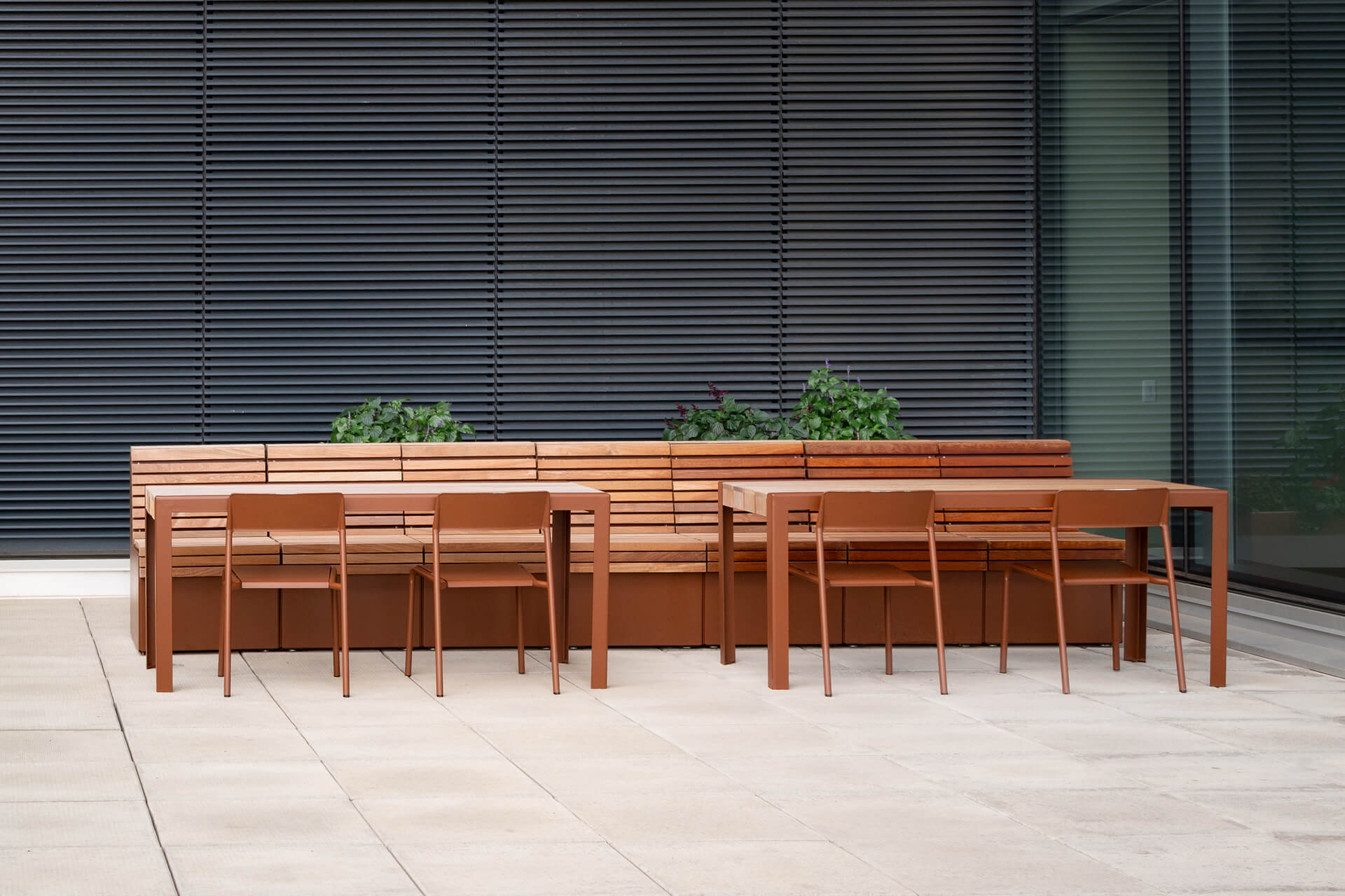 Outdoor seating area with Maglin Site Furniture, featuring two wooden 1050 tables, six matching Foro chairs, and a wooden Pixel bench with built-in planters on a tiled patio near a modern building.