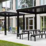 Outdoor seating area with black metal pergolas, site furniture like picnic tables, chairs, and benches in front of a modern building with large windows, thoughtfully designed for optimal space planning.