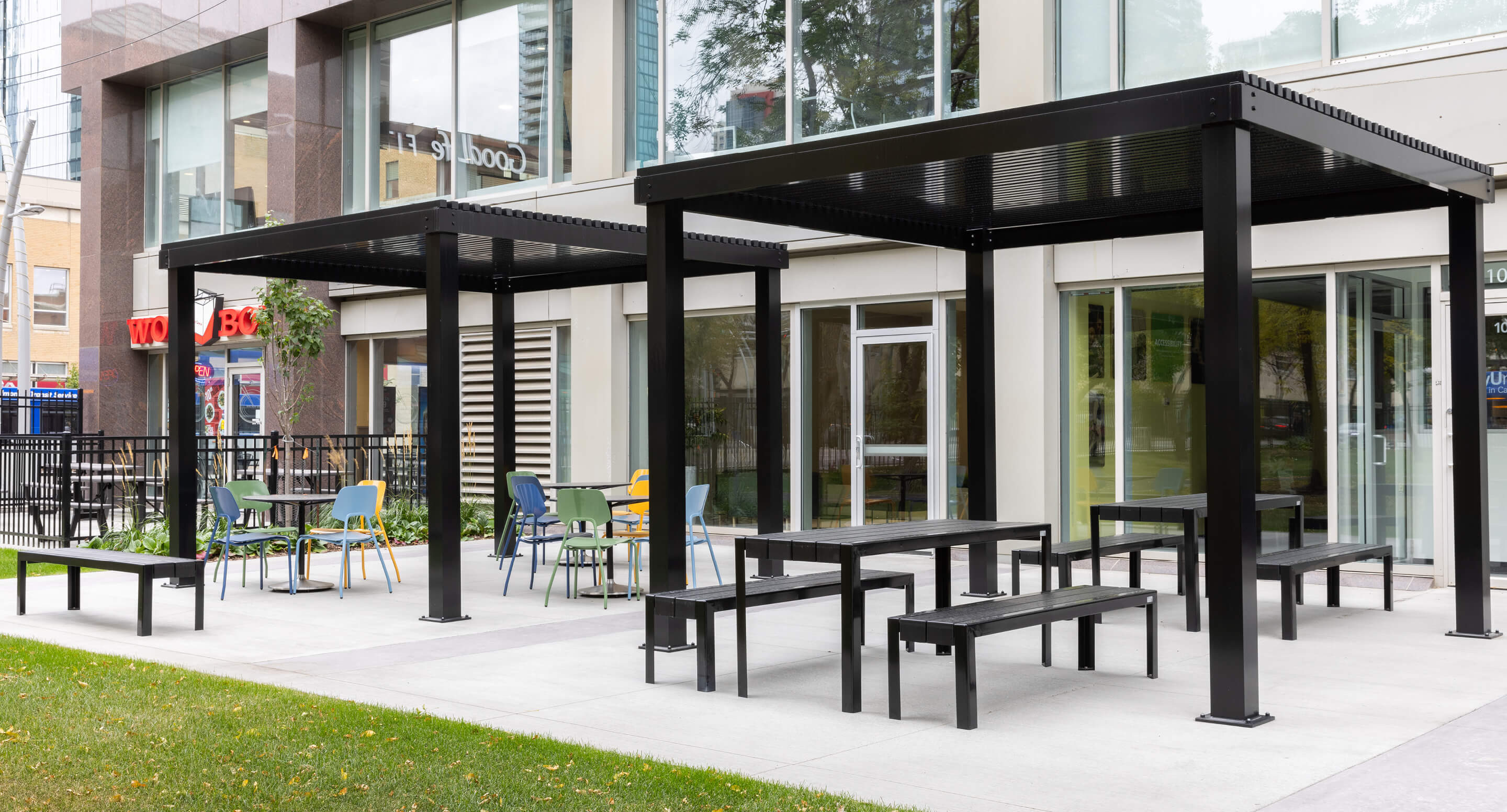 Outdoor seating area with black metal pergolas, site furniture like picnic tables, chairs, and benches in front of a modern building with large windows, thoughtfully designed for optimal space planning.