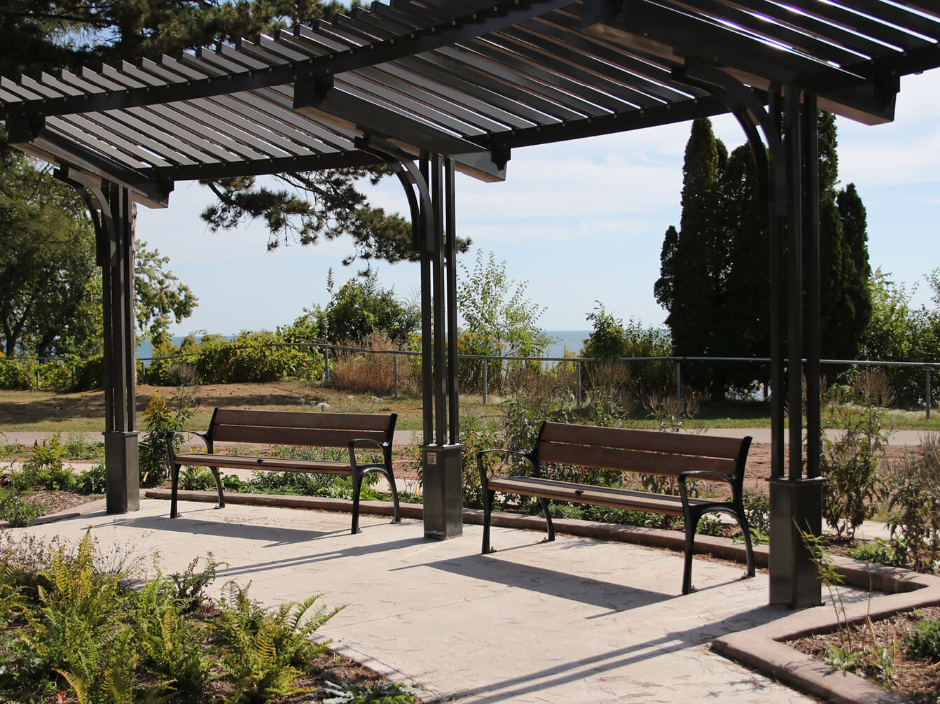 Two empty wooden benches, part of the site furniture, sit under a metal pergola in a park, with plants and trees in the background on a sunny day.