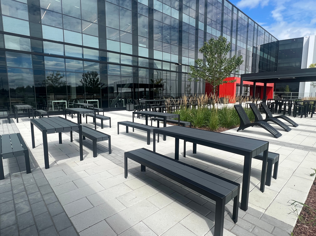 Outdoor seating area with black site furniture—tables, benches, and lounge chairs—amid landscaping, thoughtfully arranged through expert space planning next to a modern glass office building under a partly cloudy sky.