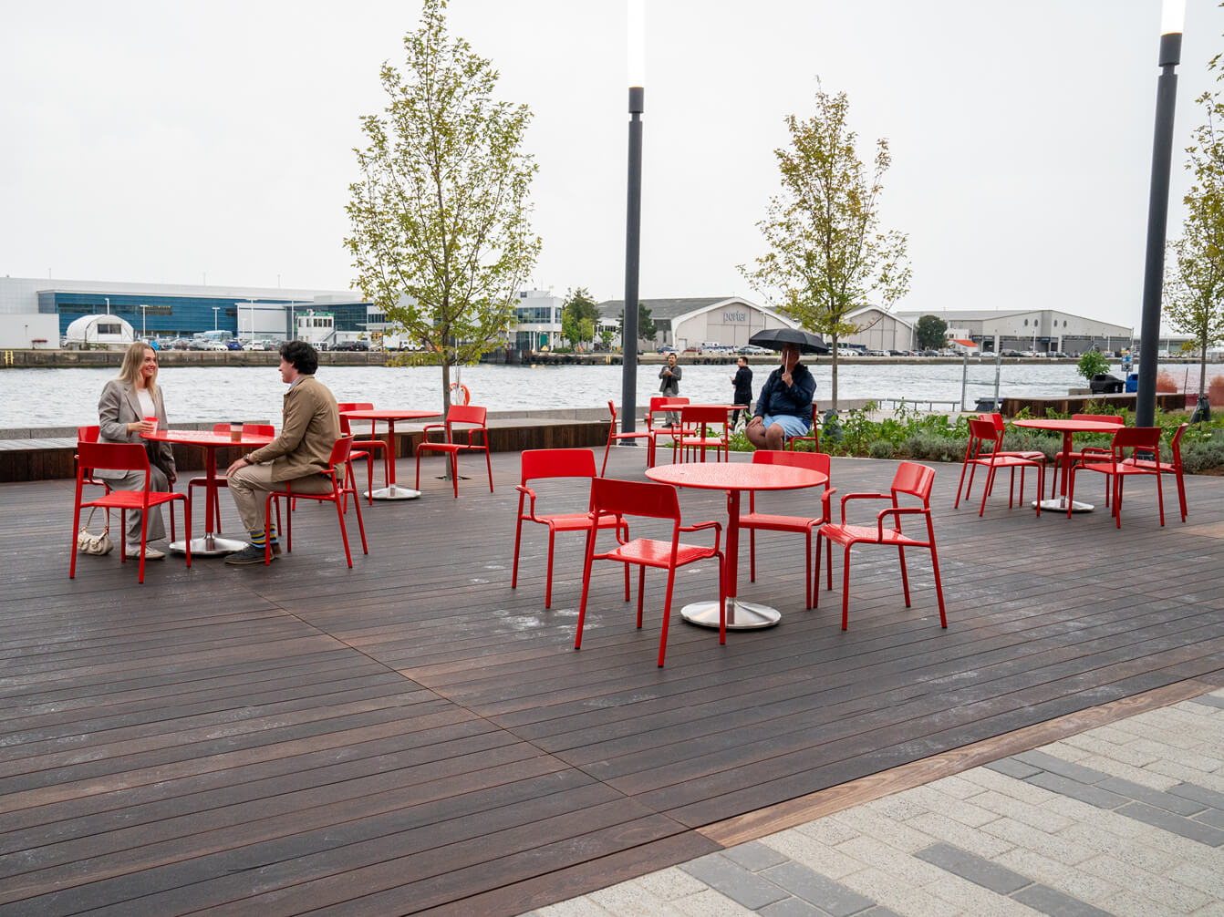 People sit at red site furniture—tables and chairs—on a wooden deck near a waterfront. Trees and a few buildings are visible in the background, enhancing thoughtful space planning in this scenic setting.