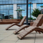 Trois chaises longues en bois sont disposées sur une terrasse extérieure carrelée, avec des bancs, des parasols verts et un bâtiment moderne en verre en arrière-plan.