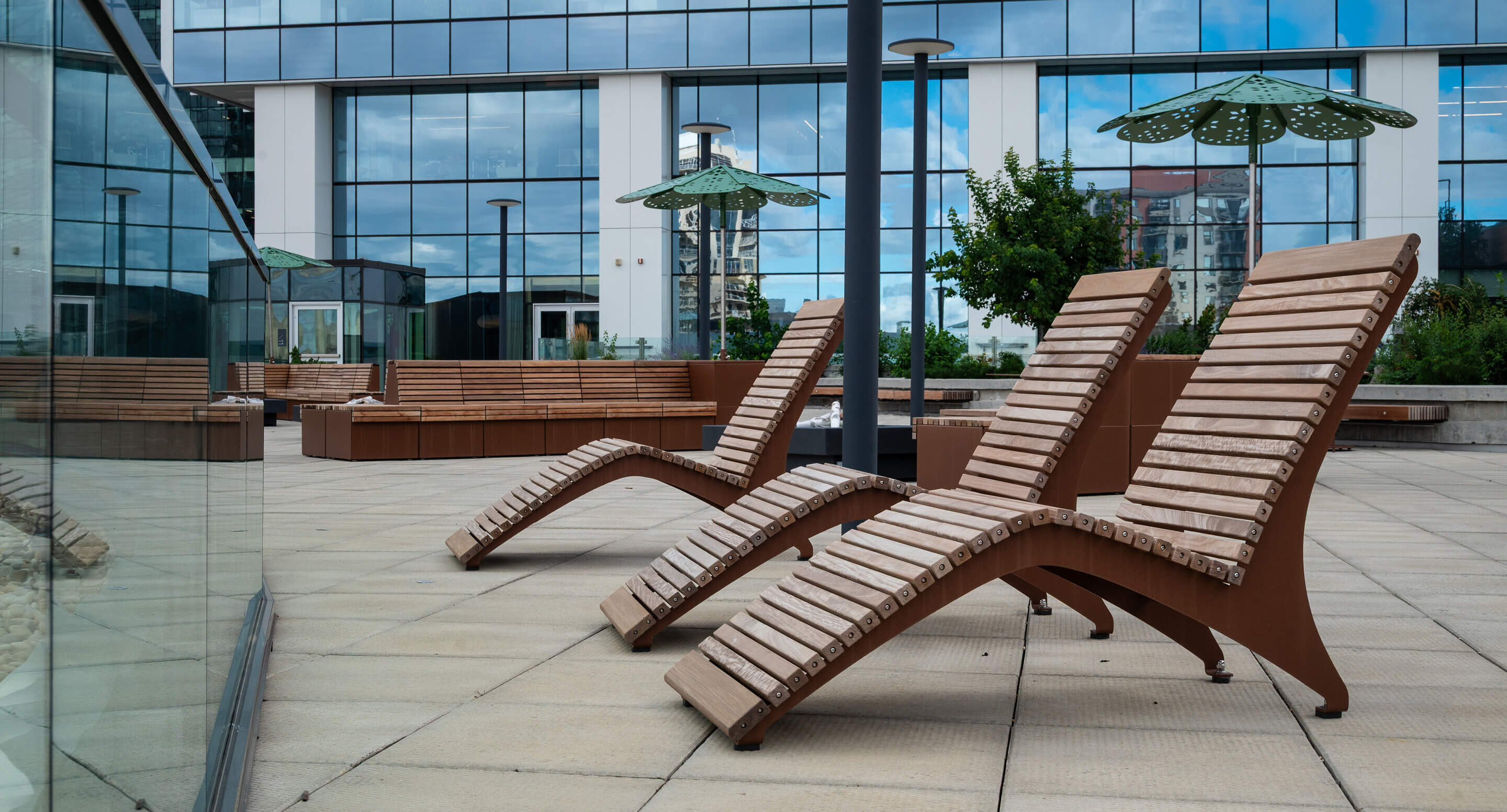 Trois chaises longues en bois sont disposées sur une terrasse extérieure carrelée, avec des bancs, des parasols verts et un bâtiment moderne en verre en arrière-plan.